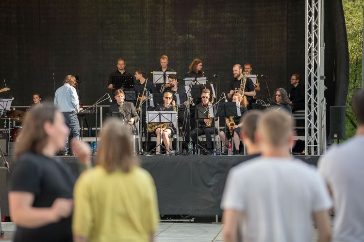 Im Vordergrund stehen Leute und hören der Unibigband zu.